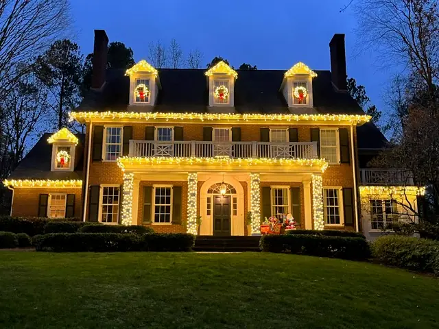 Warm lights outline the dormers, porch and windows of a brick home at dusk.