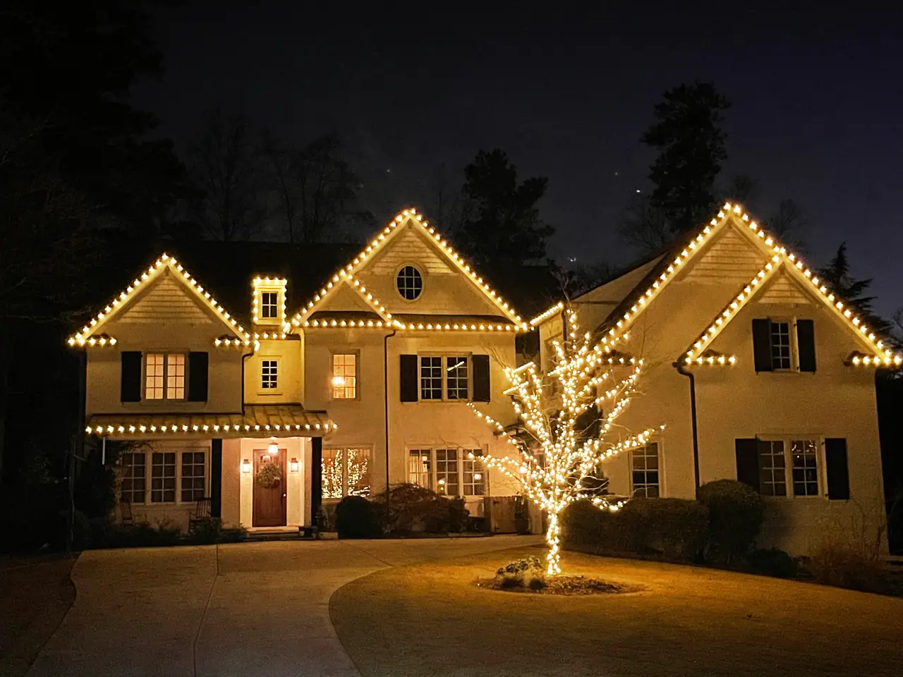 Warm white bulbs outline a home’s gables beside a tree wrapped in lights.